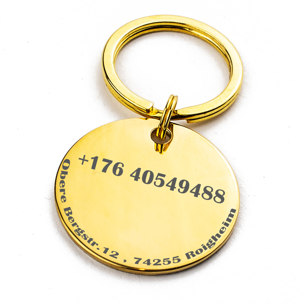 Ein goldener Schlüsselanhänger mit einem runden Anhänger, auf dem eine Telefonnummer und eine Adresse in schwarzer Schrift eingraviert sind. Es ist auf einem weißen Hintergrund fotografiert.